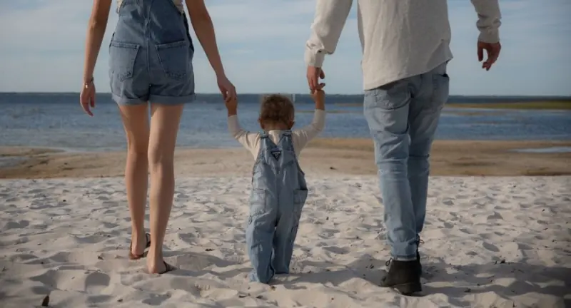 Un enfant marche au bord de l’eau en tenant la main de ses parents
